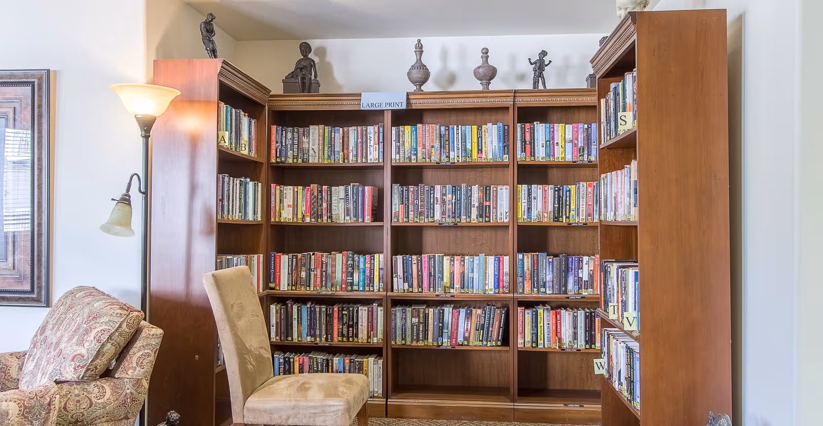 A cozy library corner in a senior living facility with wooden bookshelves filled with books, a beige upholstered chair, a patterned armchair, and a floor lamp. The bookshelves are labeled with letters and a sign indicating 'LARGE PRINT' books. Decorative statues are placed on top of the shelves.