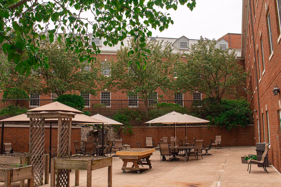 Outdoor courtyard area with patio tables, chairs, umbrellas, and wooden planters, surrounded by brick buildings and trees with green leaves.