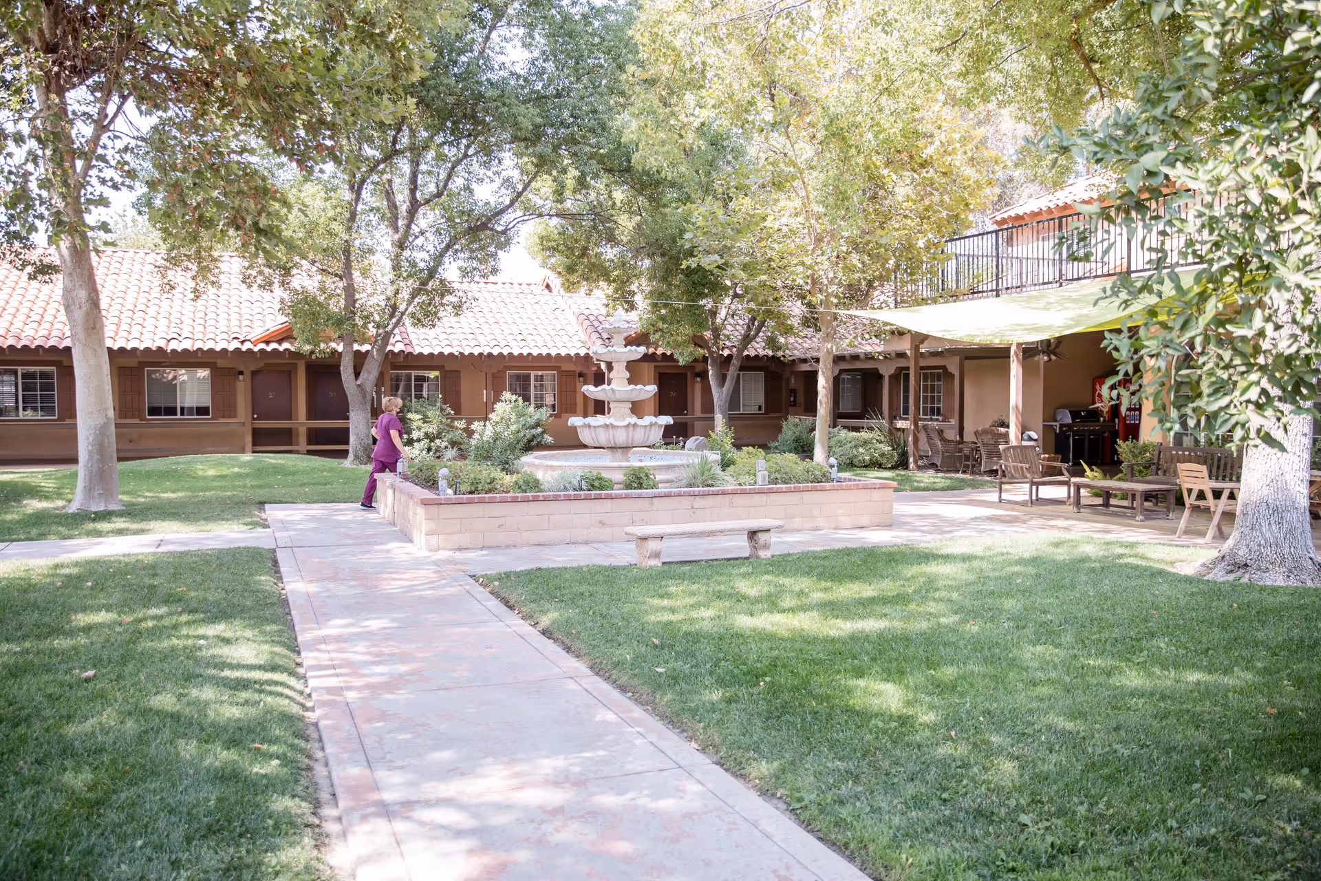 Outdoor courtyard area of Hacienda Senior Living with a central multi-tiered fountain surrounded by a low brick wall. There are trees providing shade, green grass, a paved walkway, and outdoor seating under a canopy. A person in purple clothing is standing near the fountain.