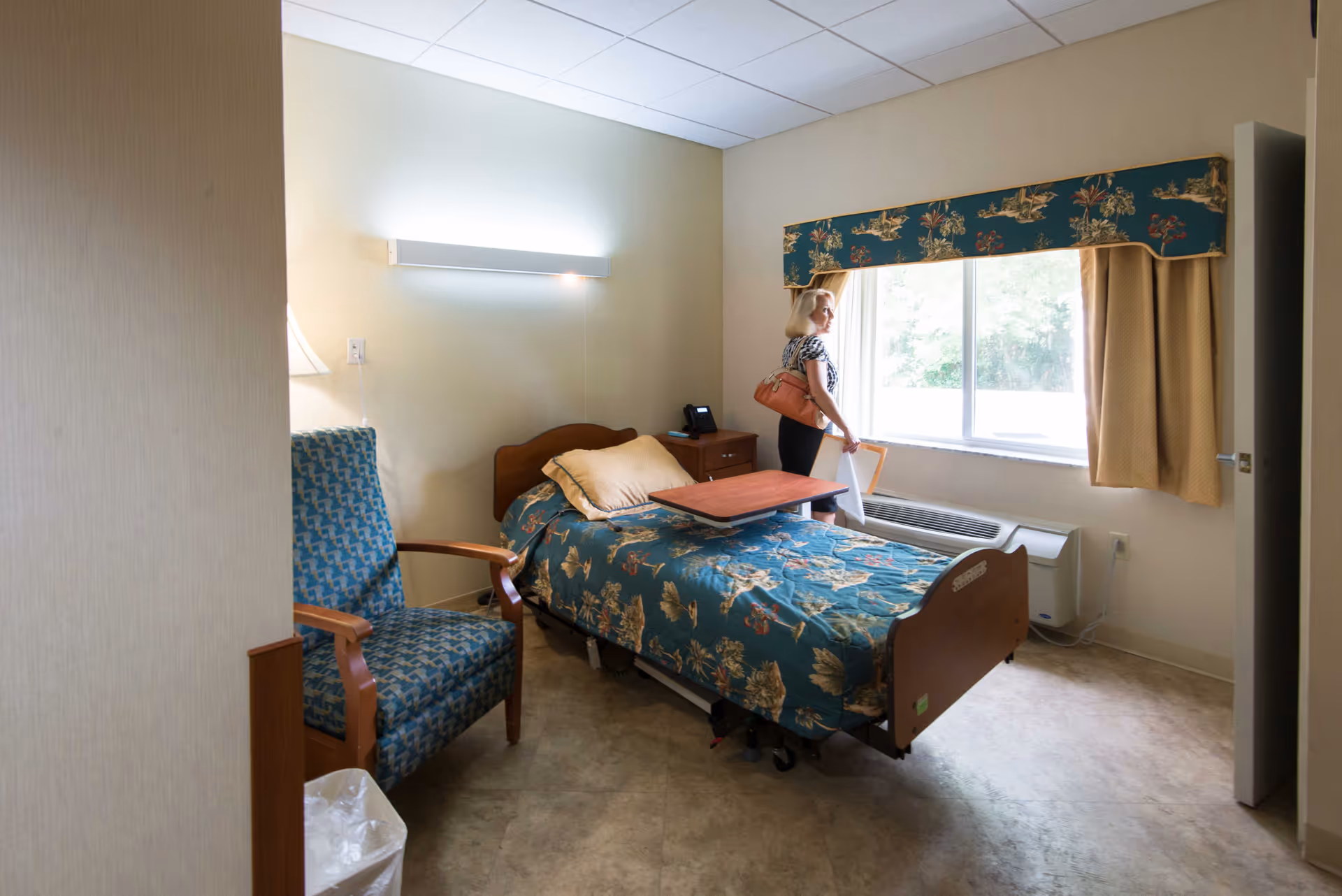 Senior living bedroom with floral bedspread, blue chair, and a resident by the window.