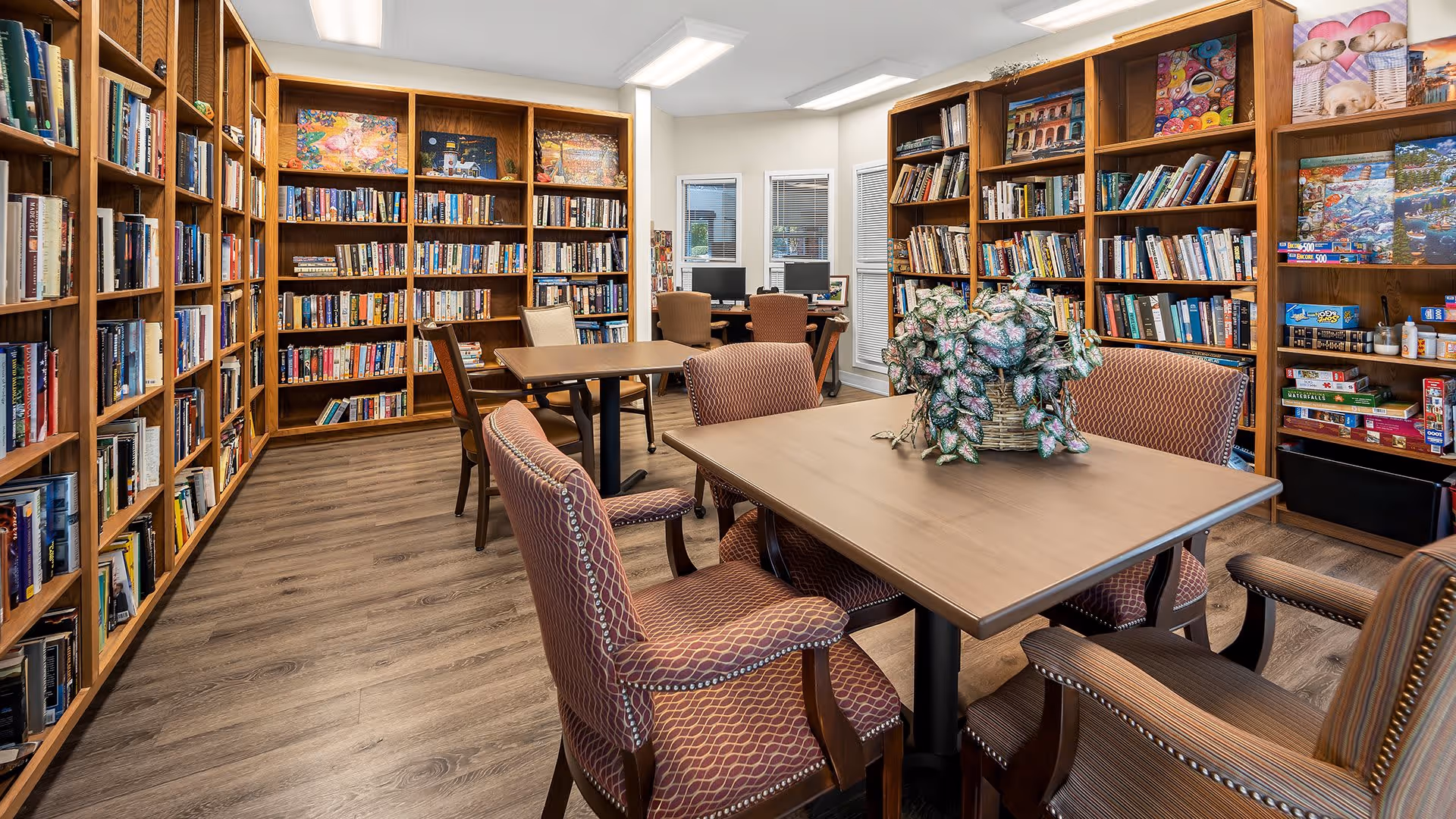A cozy library room with wooden bookshelves filled with books and board games lining the walls. There are two tables with cushioned chairs around them, one table has a decorative plant centerpiece. In the background, there are two computer stations with chairs near windows.