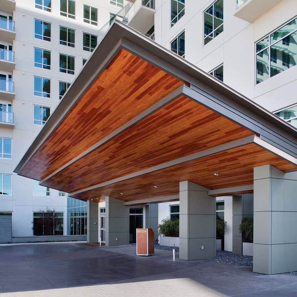 Main entrance canopy of a modern multi-story building with a wood-paneled ceiling and large supporting pillars.