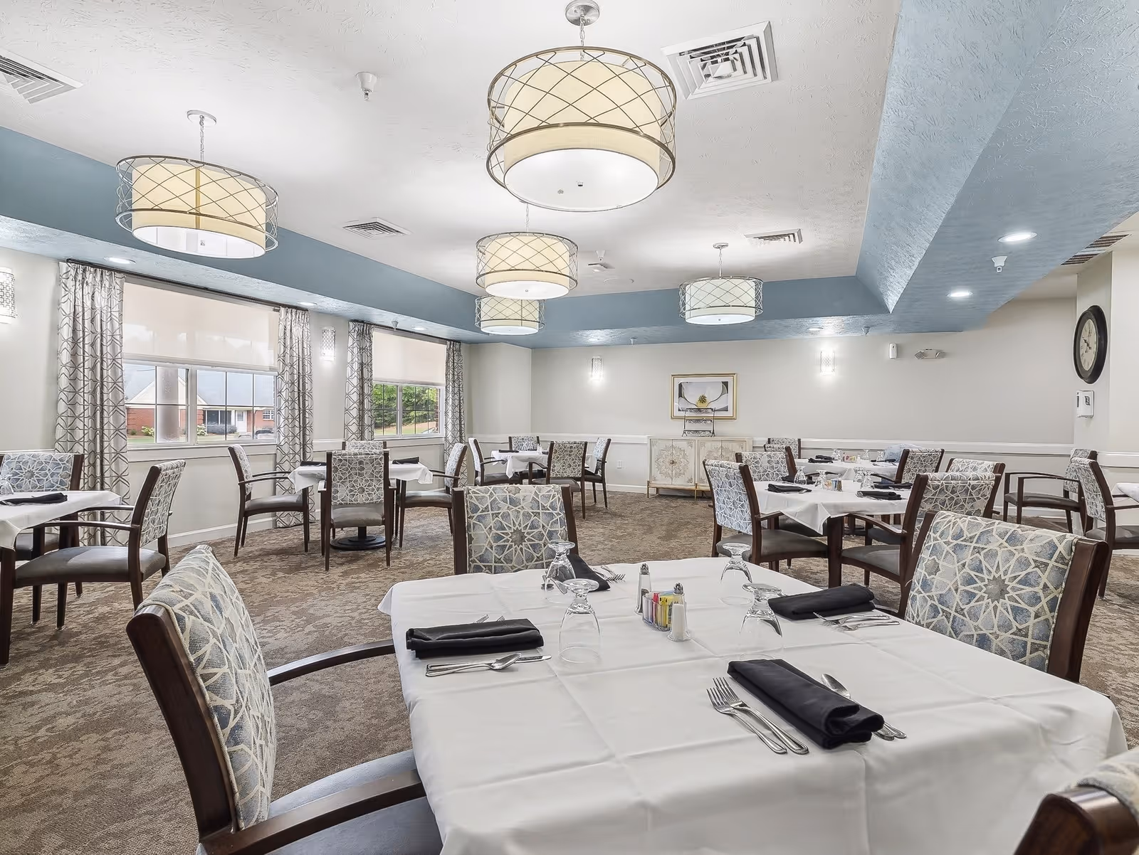 A bright and spacious dining room in a senior living facility with multiple tables covered in white tablecloths, each set with silverware, glasses, and black napkins. The room features patterned upholstered chairs, large windows with patterned curtains, and modern ceiling lights.