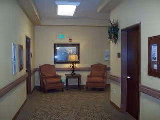 A hallway in a senior living facility with two upholstered armchairs and a small table with a lamp between them against the far wall. There is a framed picture above the chairs, a bulletin board on the right wall, and a plant hanging near a door. The walls are painted beige with a brown chair rail, and the floor is carpeted.