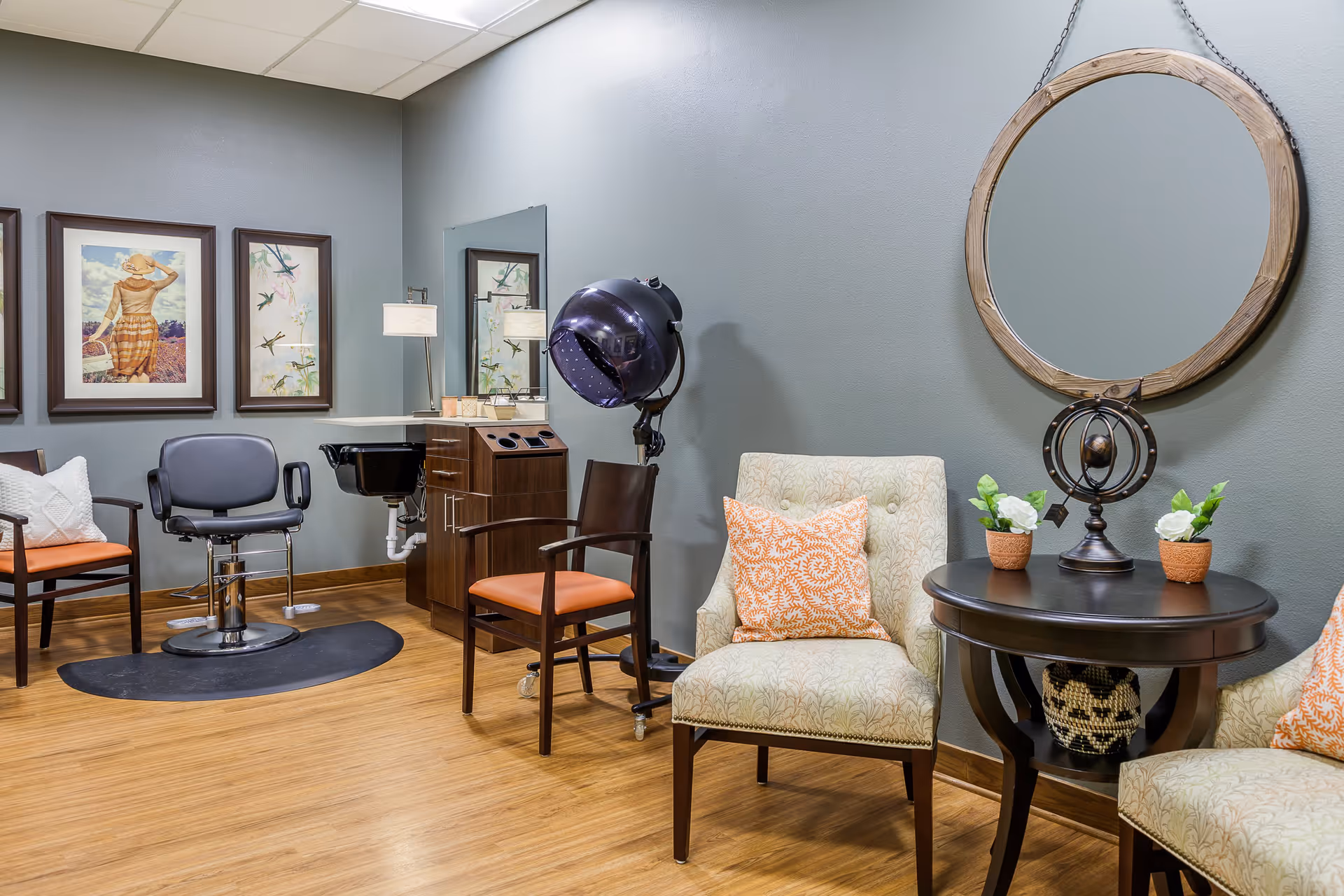 Interior of a salon area in a senior living facility with a hair styling chair, a hair dryer, a sink, two armchairs with orange patterned pillows, a round wooden table with decorative items, and framed artwork on the wall.