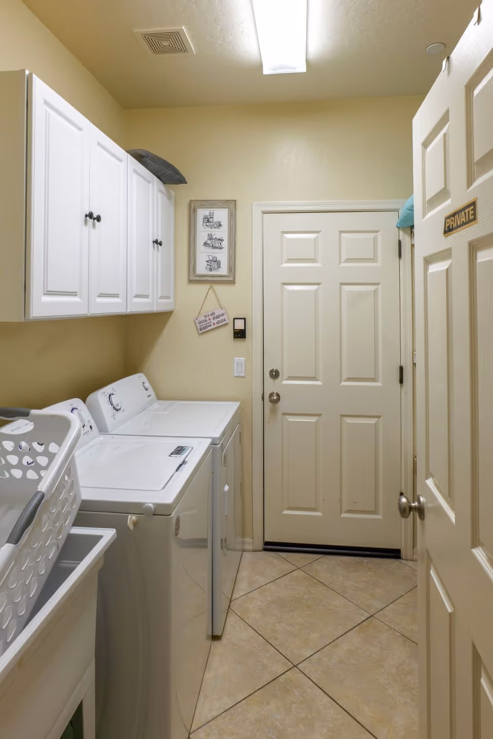 Laundry room with a white washing machine and dryer side by side, white cabinets mounted on the wall above them, a laundry basket on a white utility sink, beige tiled floor, and a closed door with a 'PRIVATE' sign.