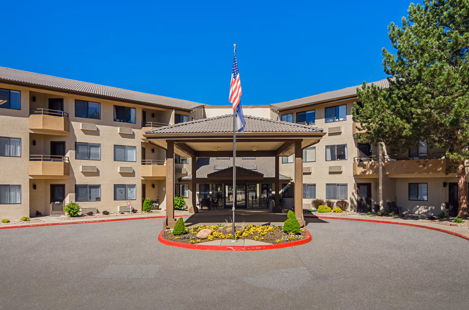Front exterior view of Solista Longmont facility with a covered entrance, American flag, and landscaped flower bed in front. The building is three stories tall with multiple windows and balconies, under a clear blue sky.