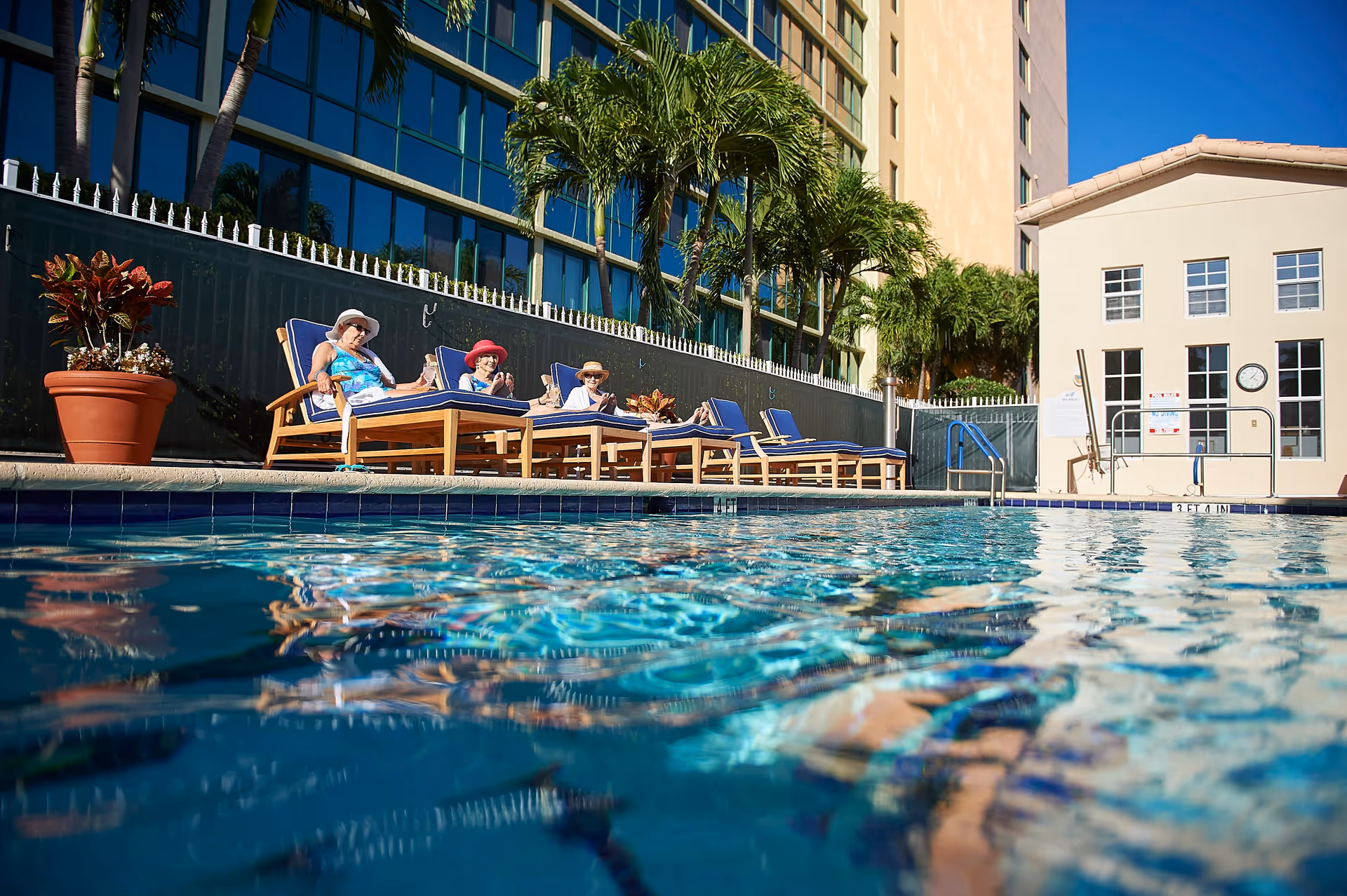 Three elderly women wearing sun hats relaxing on blue cushioned lounge chairs beside a swimming pool at a senior living facility, with palm trees and a multi-story building in the background under a clear blue sky.