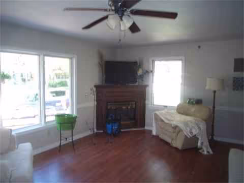 Bright living room in a senior facility with fireplace, ceiling fan, TV, and seating.