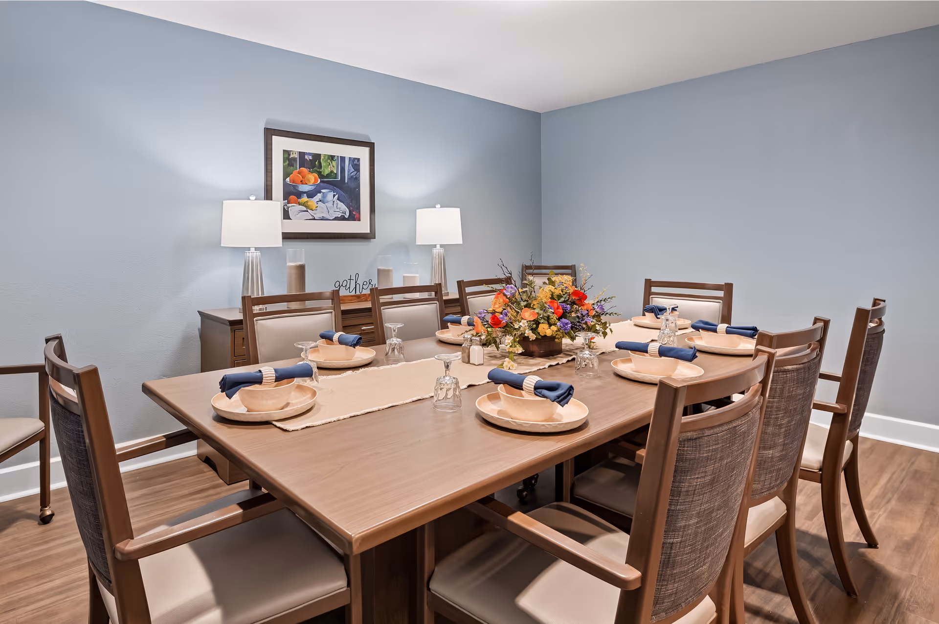 A dining room with a large wooden table set for eight people. Each place setting includes a beige plate, a bowl, a glass turned upside down, and a blue cloth napkin with a napkin ring. A floral centerpiece decorates the middle of the table. The room has light blue walls, wood flooring, a sideboard with two lamps, and a framed painting of fruit on the wall.