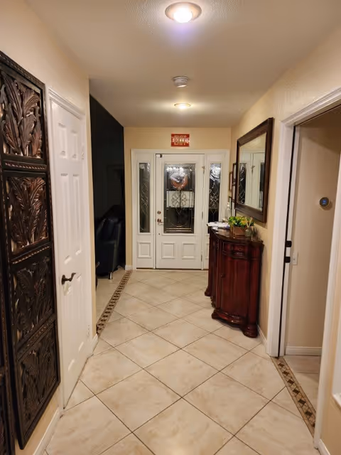 A well-lit hallway with beige tiled floor and cream-colored walls. On the left side, there is a decorative wooden panel and a white door. On the right side, there is a wooden console table with plants and a large mirror above it. At the end of the hallway, there is a white front door with decorative glass panels and sidelights.