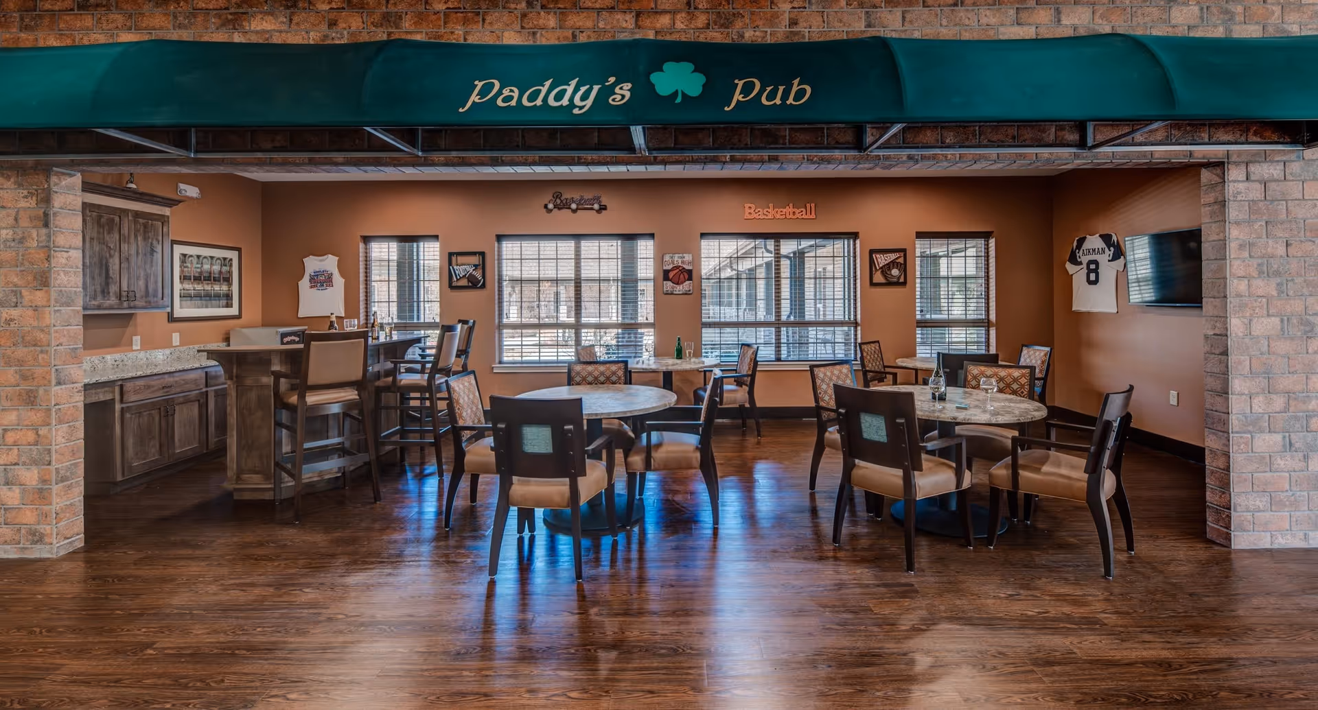 Interior view of Paddy's Pub, a cozy room with wooden floors, round tables with chairs, a bar counter with high stools, sports-themed decorations on the walls including jerseys and framed pictures, and large windows letting in natural light.