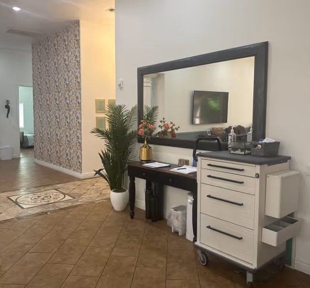A tiled hallway or reception area with a console table under a large mirror, a potted plant, wall-mounted TV reflected in the mirror, and a wheeled storage cart.