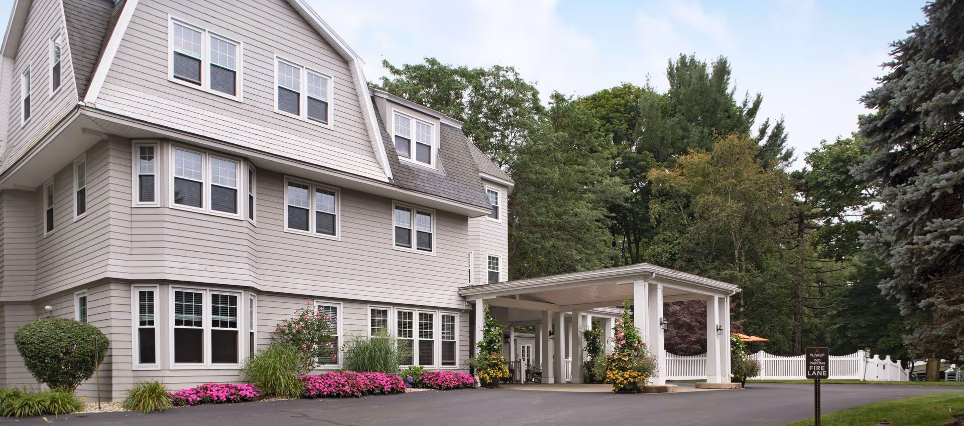 Exterior view of a multi-story senior living facility building with gray siding and numerous windows. The entrance features a covered drop-off area adorned with hanging flower baskets. The surrounding area includes well-maintained landscaping with bushes, pink flowers, and tall trees in the background. A sign near the driveway reads 'Sunrise No Parking Fire Lane.'