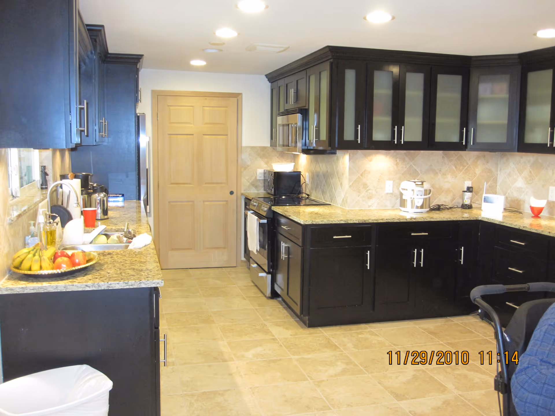 A kitchen with dark wood cabinets, granite countertops, and beige tiled floor and backsplash. The kitchen features a stove, microwave, sink, and various kitchen appliances and items including a fruit plate with bananas and apples. There is a wooden door at the end of the kitchen and recessed ceiling lights.