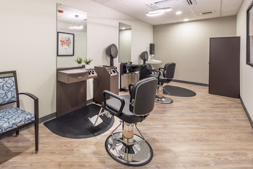 Interior view of a salon area in a senior living facility with two black salon chairs in front of mirrors and hair drying stations. The room has light-colored walls, wood flooring, a patterned chair on the left, a small cabinet on the right, and framed artwork on the walls.