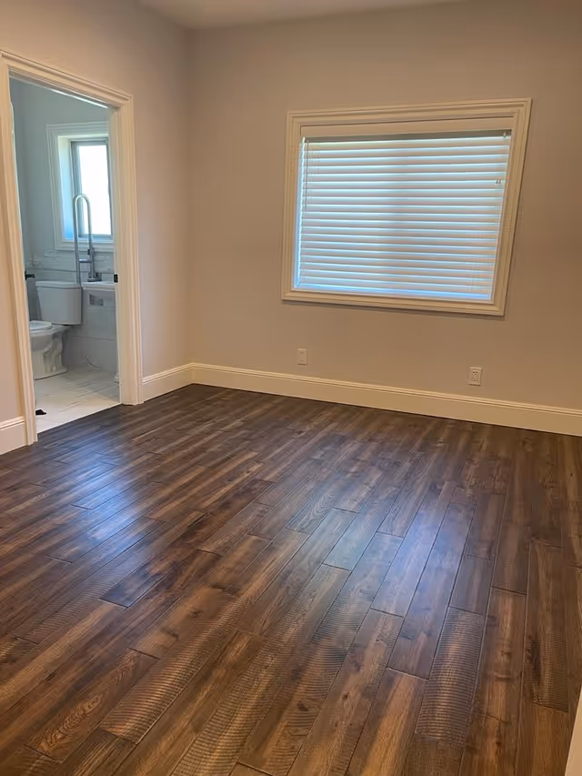 Empty room with dark hardwood floors, a window with closed blinds, and an open doorway showing a bathroom.