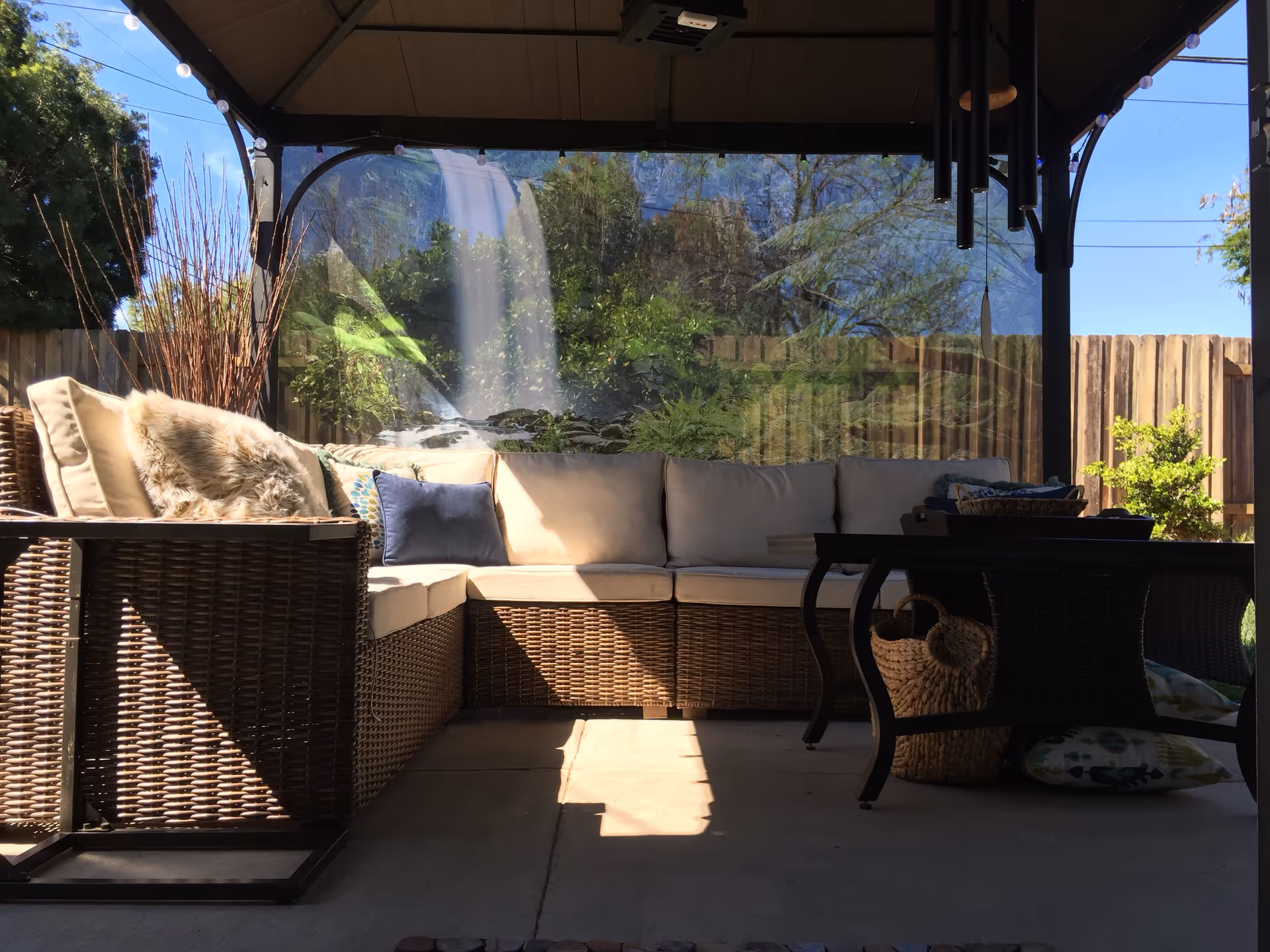 Outdoor covered patio area with a wicker sectional sofa featuring beige cushions and decorative pillows. A wicker coffee table with a basket underneath is in front of the sofa. The patio is enclosed with a transparent screen, and a wooden fence and greenery are visible outside.