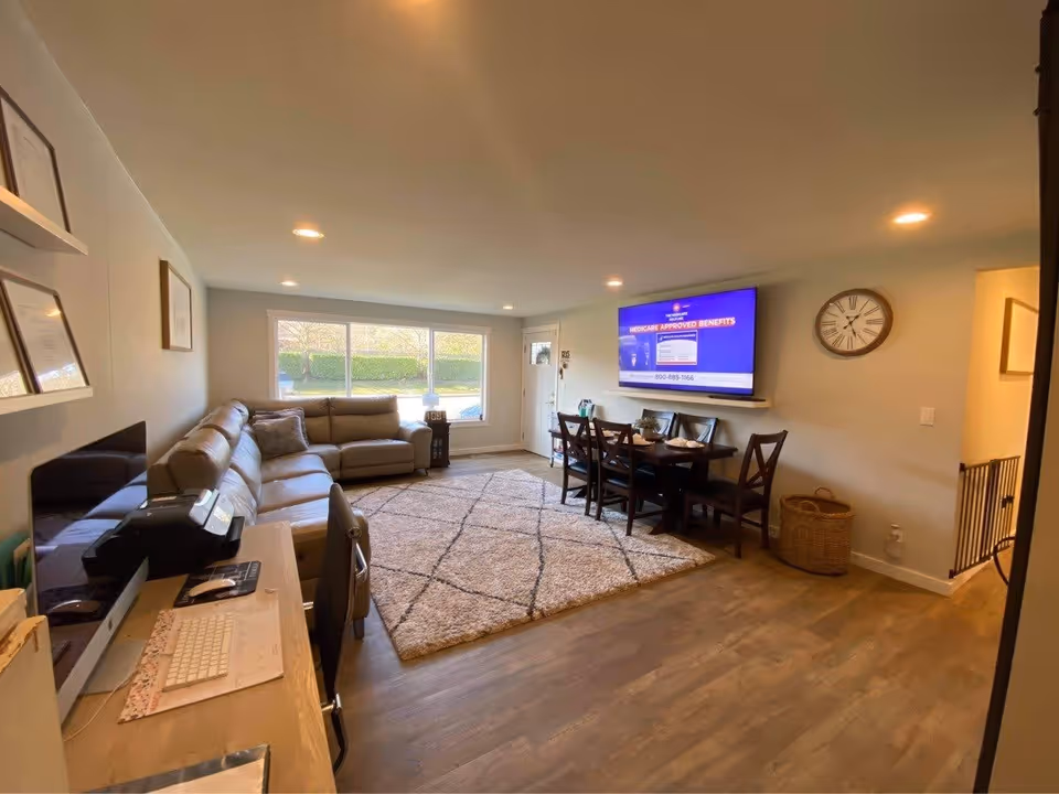 A cozy living room with a large beige sectional sofa, a patterned area rug, a wooden dining table with six chairs, a wall-mounted flat screen TV displaying Medicare approved benefits information, a clock on the wall, and a desk with a computer and office chair. Large windows let in natural light and show greenery outside.