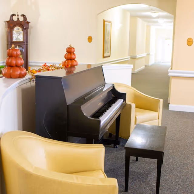 A cozy indoor seating area with a black upright piano, two yellow armchairs, and a small black table. The space is decorated with autumn-themed pumpkin ornaments and a grandfather clock is visible in the background along a well-lit hallway.