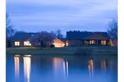 Evening view of a lakeside property with three illuminated buildings reflecting on the calm water under a cloudy sky.