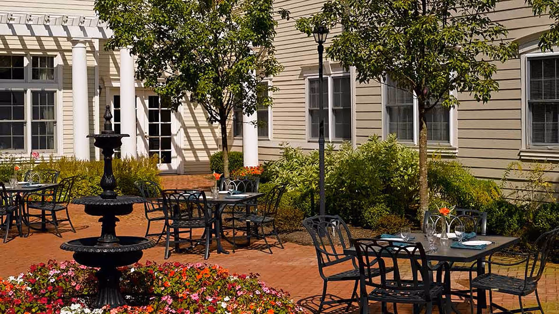 Sunny courtyard with a tiered fountain, outdoor tables and chairs surrounded by flowers and a building facade.