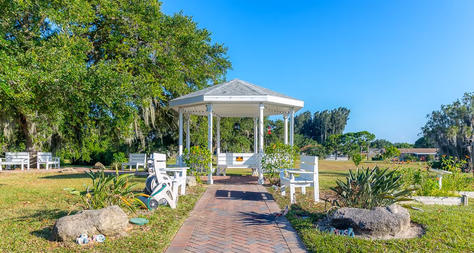 A peaceful outdoor garden area with a white gazebo at the center, surrounded by white benches and green plants. Large trees provide shade, and a brick pathway leads to the gazebo under a clear blue sky.