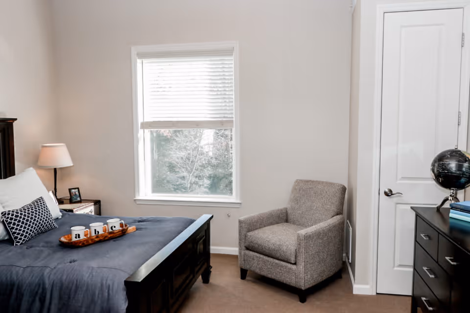 Sunlit bedroom with a made bed, bedside table and lamp, upholstered armchair, and dresser with a globe.