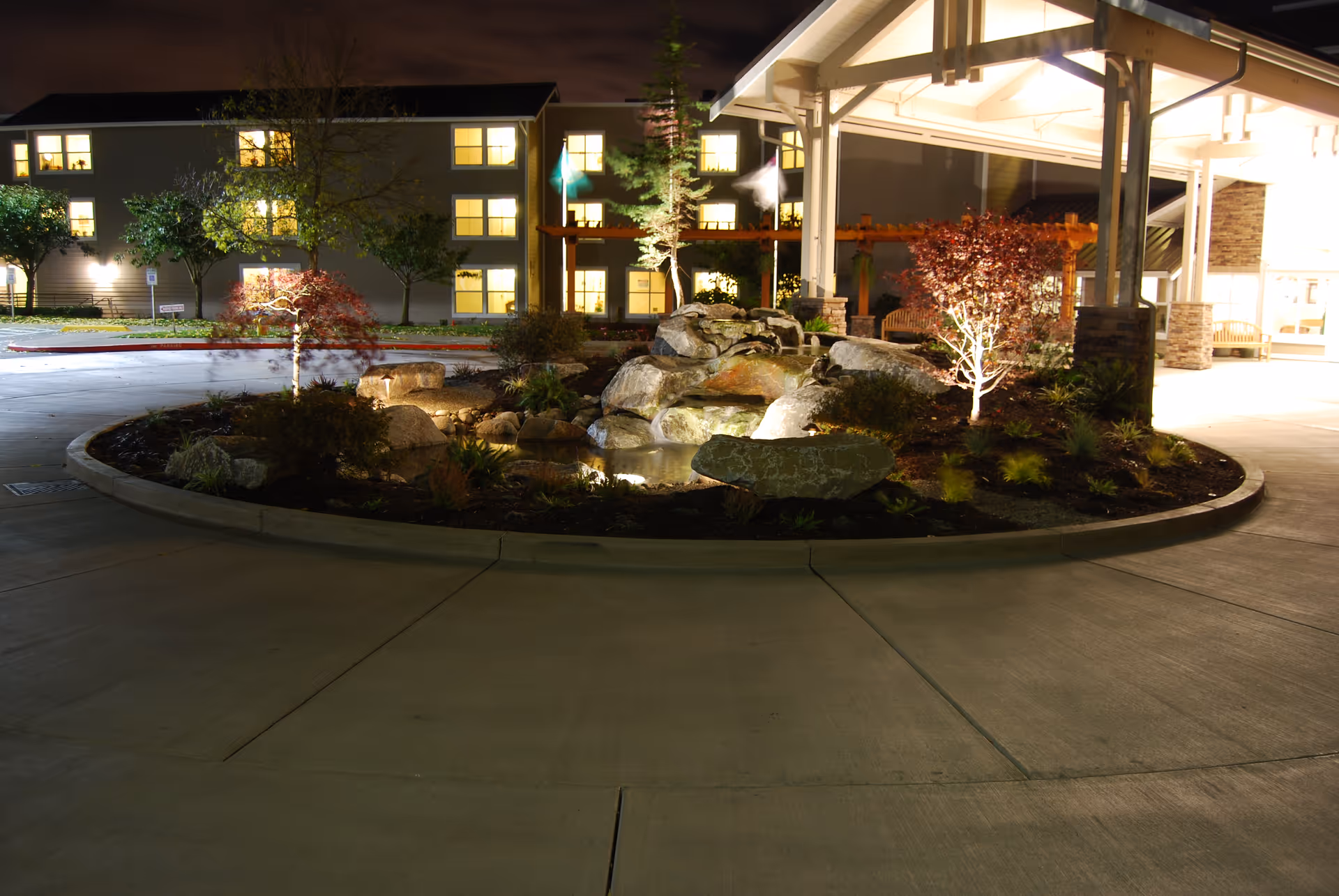 Night view of the entrance area of Crista Rehab & Skilled Care facility featuring a circular landscaped island with rocks, plants, and small trees illuminated by lights. The building with multiple lit windows is visible in the background along with a covered entrance structure.