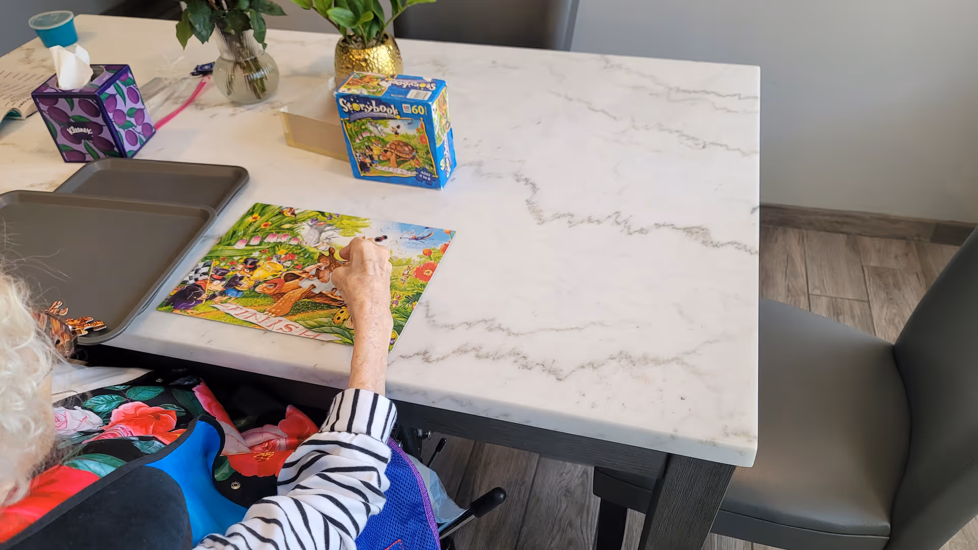 An elderly person with white hair is sitting at a marble-top table working on a colorful jigsaw puzzle. On the table are two trays, a box of tissues, a box for the puzzle, and two small vases with green plants. The floor is wooden, and there is a gray chair next to the table.