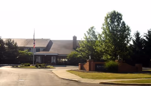 Front exterior view of Kingston Residence of Marion building with a driveway, landscaped greenery, and an American flag on a flagpole.