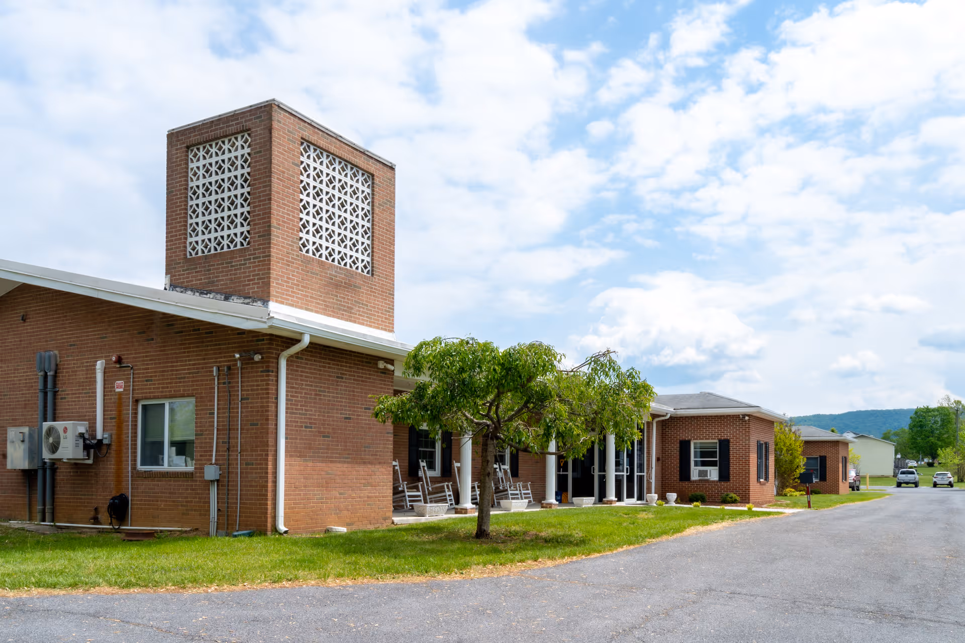 Exterior view of a single-story brick nursing home building with a distinctive brick tower featuring decorative latticework. There is a small tree and green lawn in front of the building, a paved driveway, and a partly cloudy sky overhead.