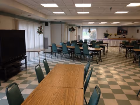 Large dining/activity room with rows of tables and green chairs, checkered tile floor, a TV on the left and a buffet table at the far end.
