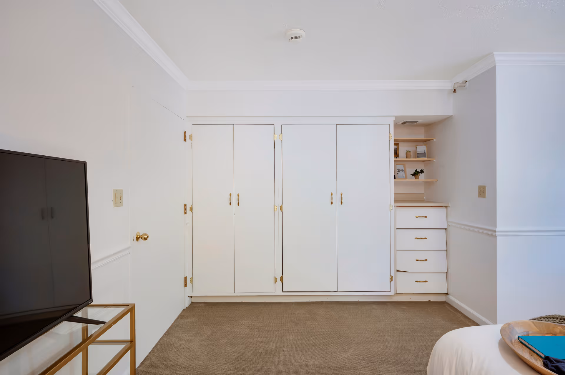 A corner of a bedroom in an assisted living facility showing white built-in closets and drawers with gold handles, a flat-screen TV on a glass and gold stand, and part of a bed with a wooden tray holding a blue book and some clothing.