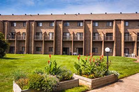Brick multi-story senior living building with balconies, a manicured lawn, and planter boxes in front.