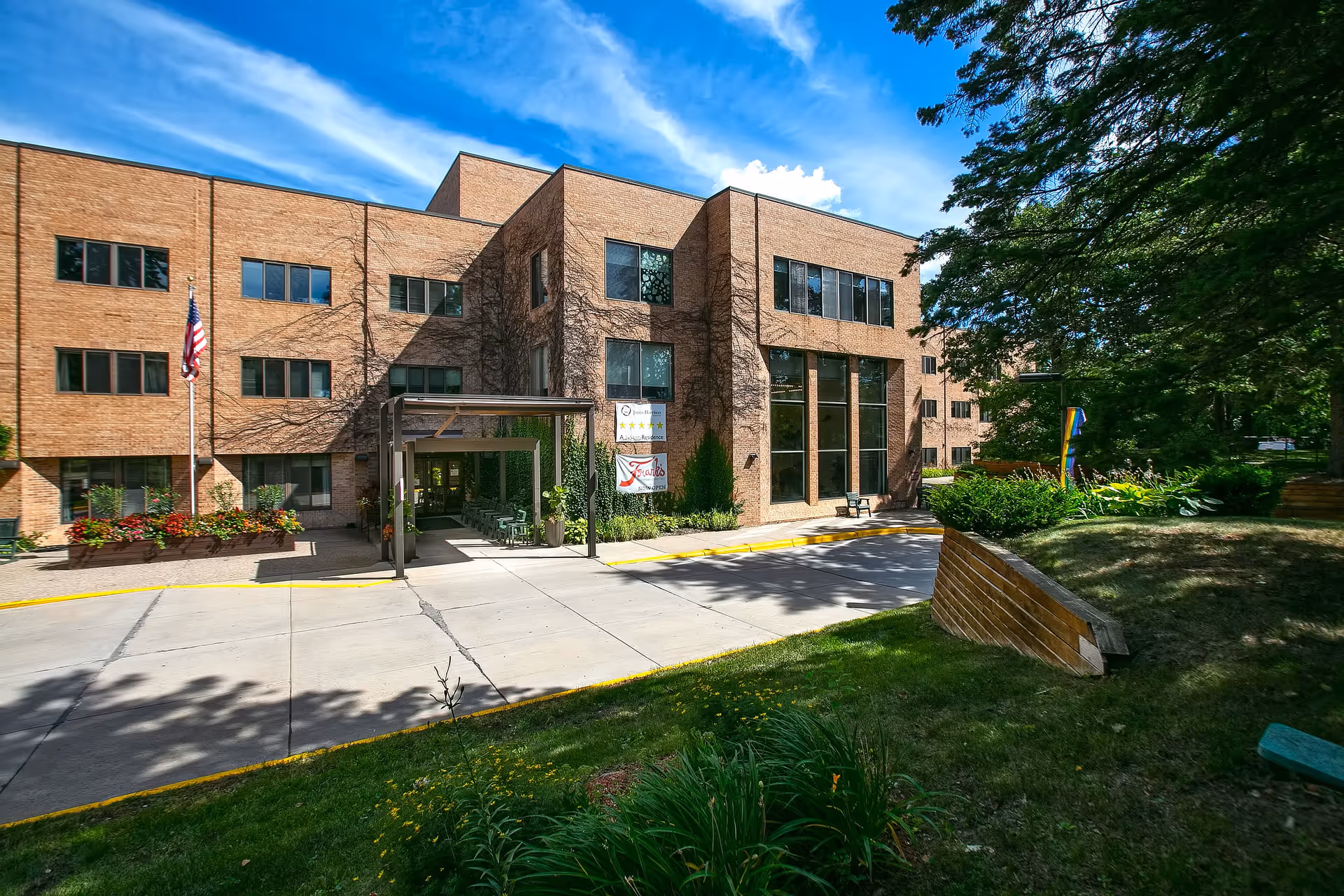 Exterior view of a three-story brick building with large windows and an entrance canopy. The building is surrounded by greenery, including trees, bushes, and flower beds. An American flag is visible near the entrance, and a sign near the door reads 'Jones-Harrison' and 'Frank's Now Open'. The sky is blue with some clouds.