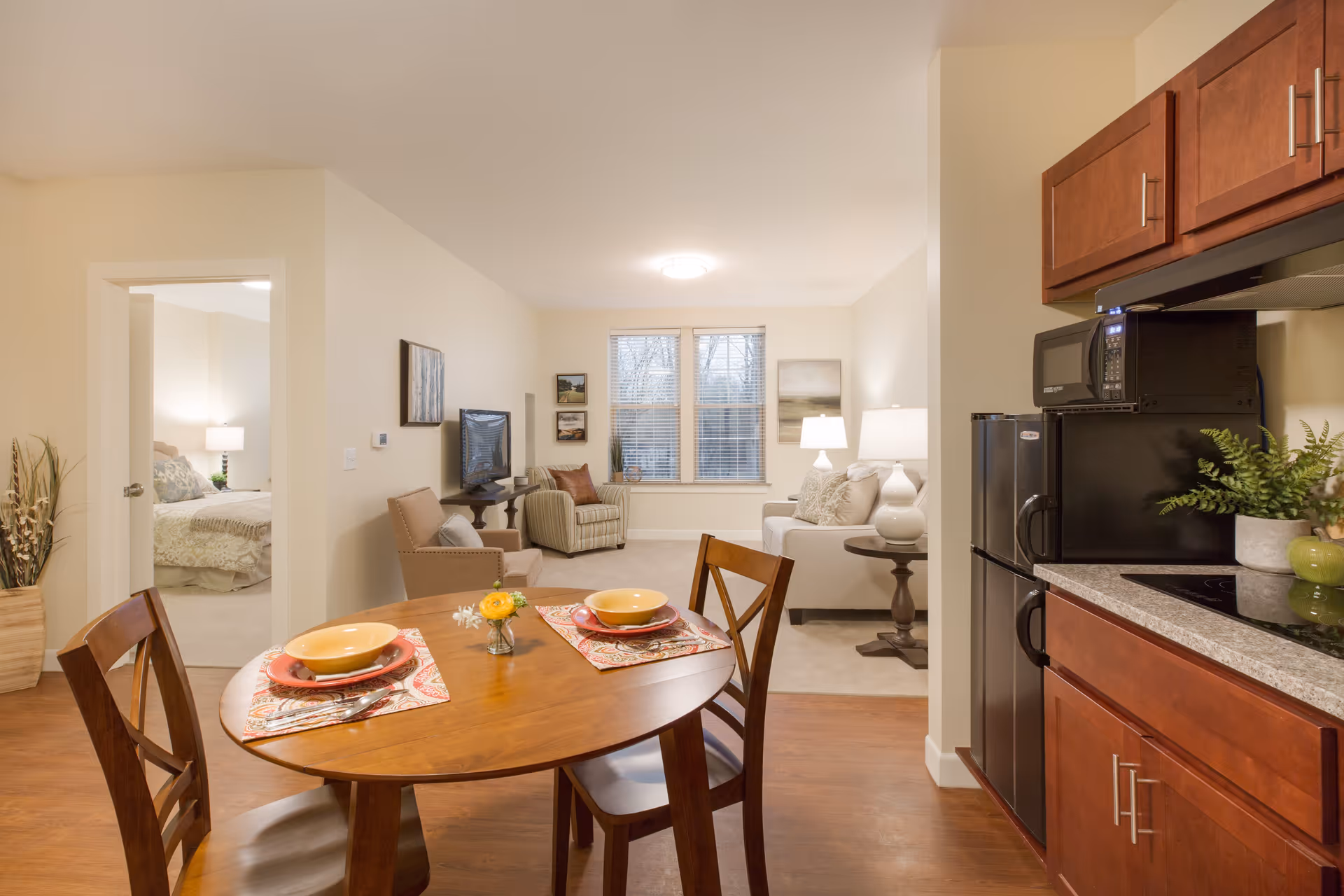 Interior view of a senior living apartment at The Residence at Orchard Grove showing a dining area with a wooden table set for two, a kitchen with wooden cabinets and appliances, and a living room with a sofa, armchair, TV, and window. A bedroom is visible through an open door on the left.