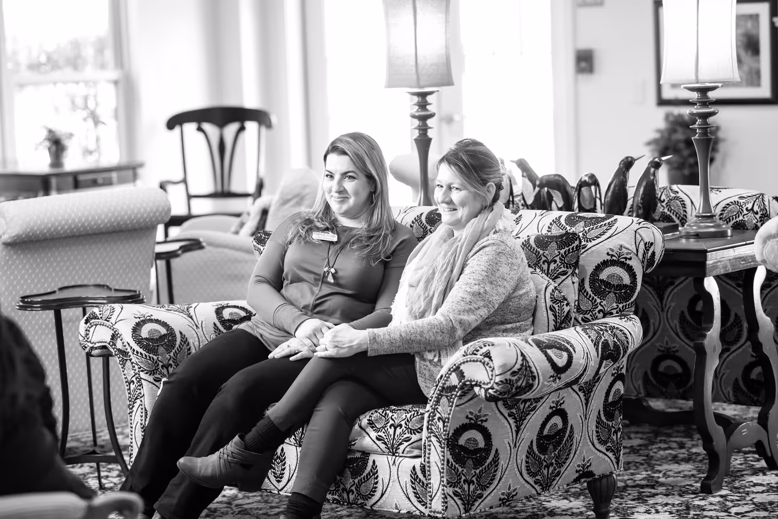 Two women sitting on a patterned sofa in a senior living lounge, smiling and holding hands.