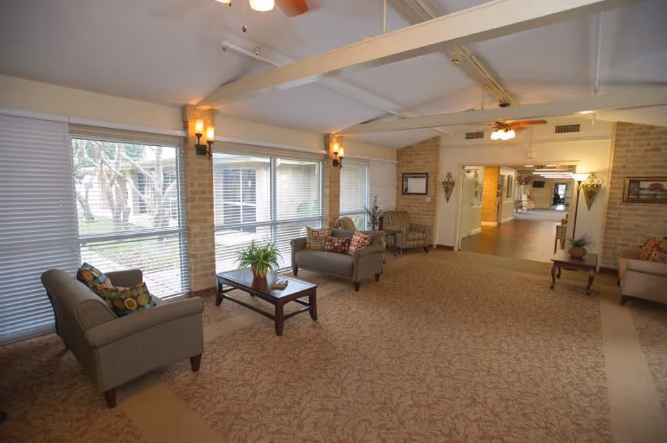 A spacious and well-lit common area in a senior living facility with large windows covered by blinds, beige carpet with a subtle pattern, and several seating arrangements including armchairs and sofas with decorative pillows. There is a wooden coffee table with a plant on it, wall sconces providing warm lighting, and ceiling fans. The room opens into a hallway leading to other parts of the facility.