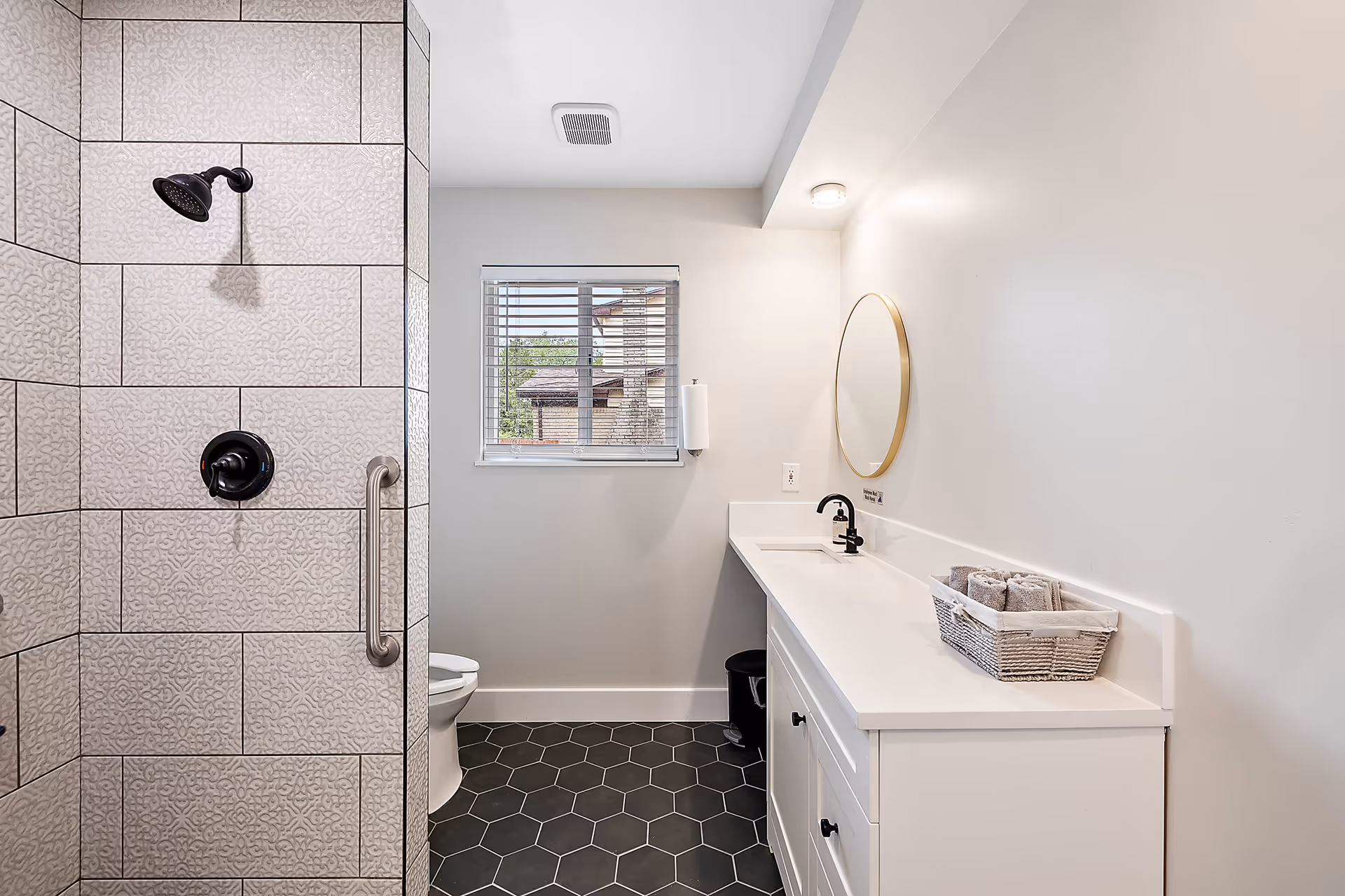 A modern bathroom featuring a tiled shower with a black showerhead and handle, a white vanity with a black faucet, a round mirror with a gold frame, a basket with rolled towels on the counter, a window with blinds, and hexagonal black floor tiles.