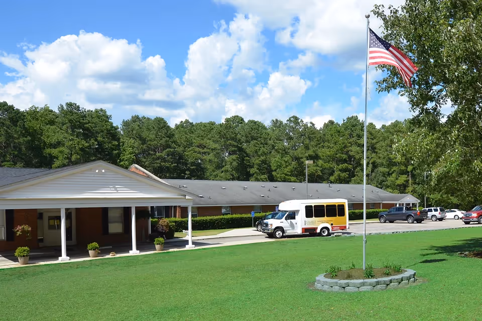 Single story retirement center entrance with lawn, flagpole, bus, and parked cars in a suburban setting.