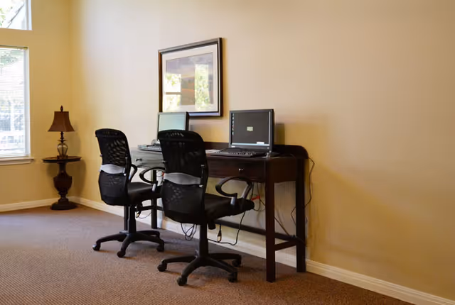 A small computer workstation area with two black office chairs and two desktop computers on a dark wooden desk against a beige wall. A framed picture hangs above the desk, and a decorative lamp on a small table is positioned near a window with blinds partially open, letting in natural light.
