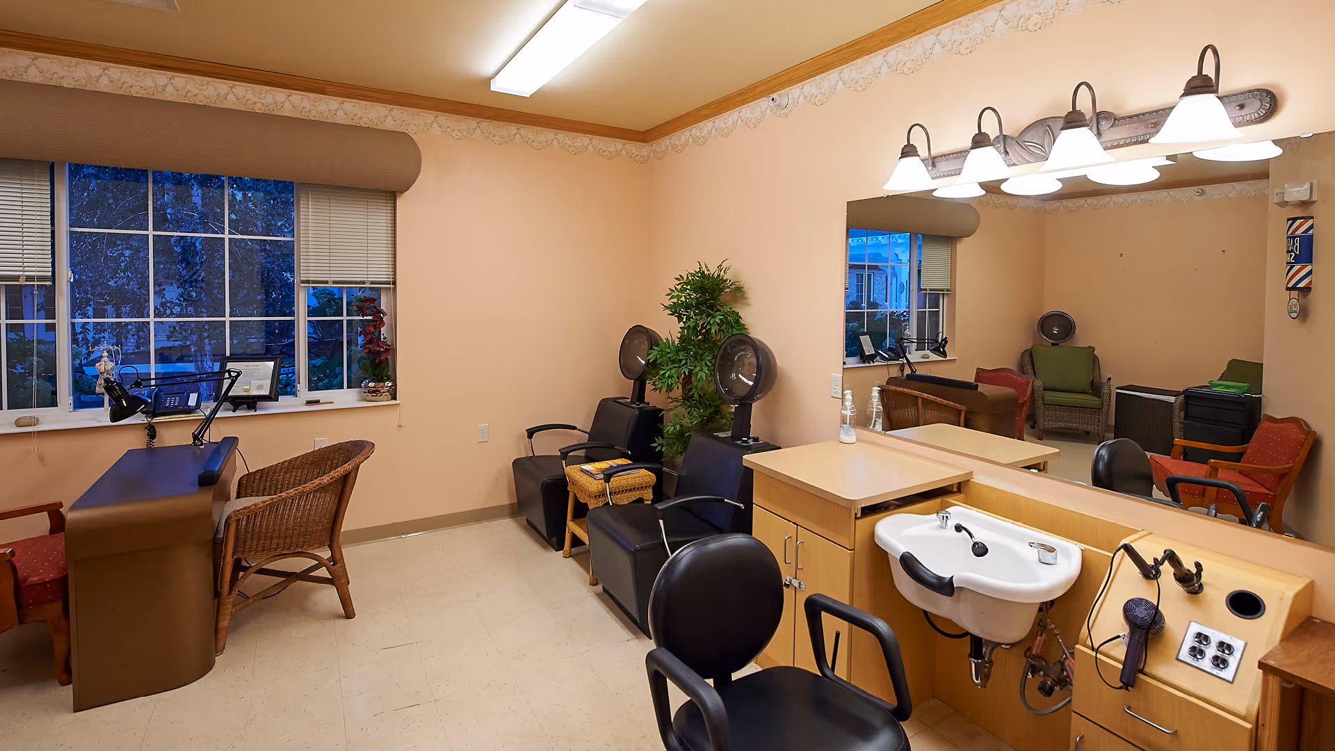 Interior of a salon room in a senior living facility with two black salon chairs and hair dryers, a large mirror with lights above it, a small sink with a hairdryer attached, a desk with a wicker chair, and a window with blinds showing outdoor trees.