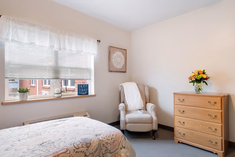 A cozy bedroom with a bed covered in a patterned quilt, a window with blinds and a white valance, a beige armchair with a white throw blanket, a wooden dresser with a vase of colorful flowers, and a framed decorative artwork on the wall.