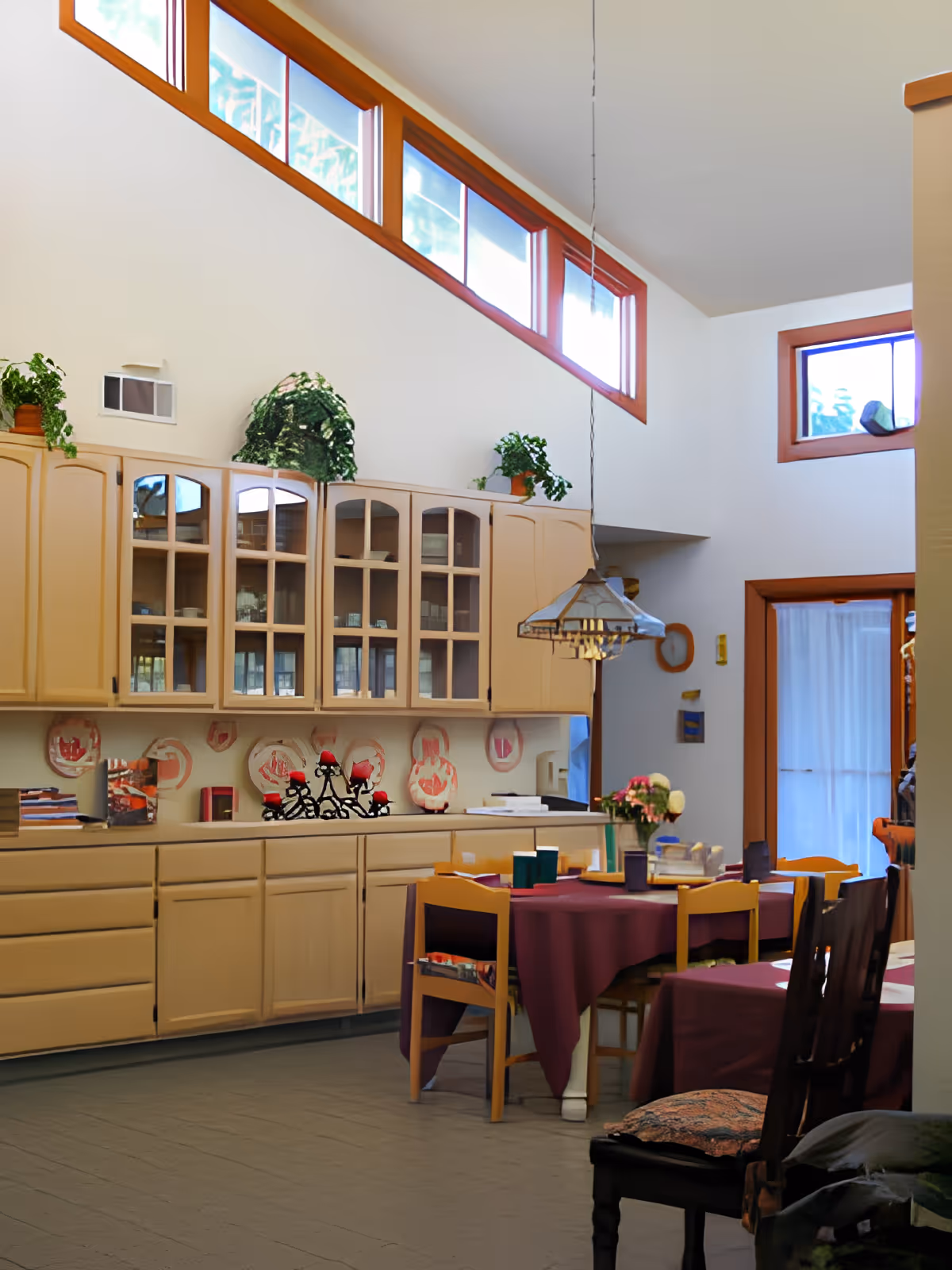 Interior view of a dining area with a table covered in a purple tablecloth surrounded by wooden chairs. Behind the table is a long counter with beige cabinets and glass-front upper cabinets. There are plants on top of the cabinets and decorative plates on the wall. High windows near the ceiling allow natural light into the room.