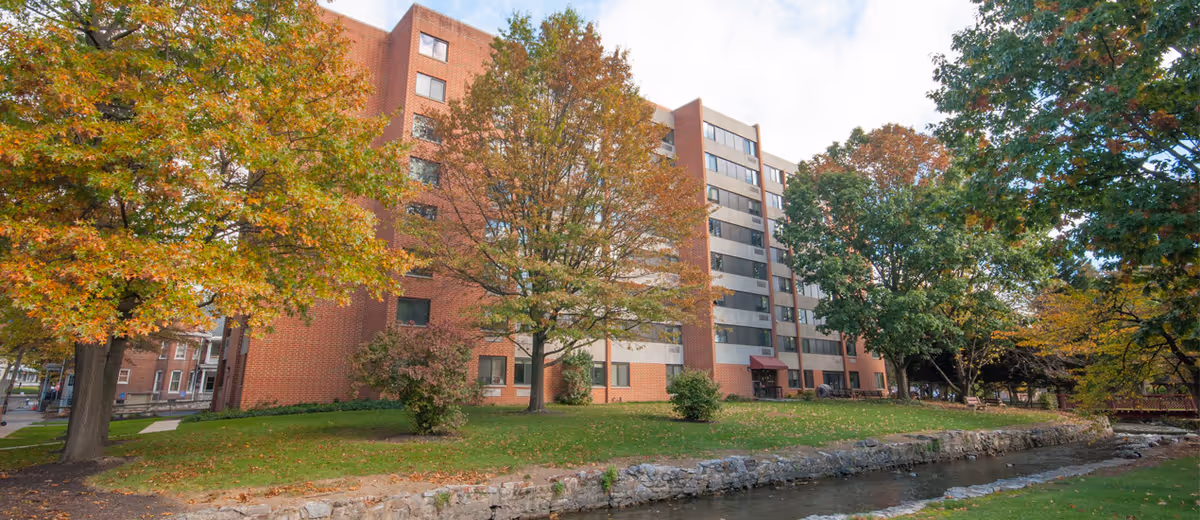 Exterior view of a multi-story brick residential building named The Tower at Falling Spring, surrounded by trees with autumn foliage and a small stream running alongside a grassy area in the foreground.