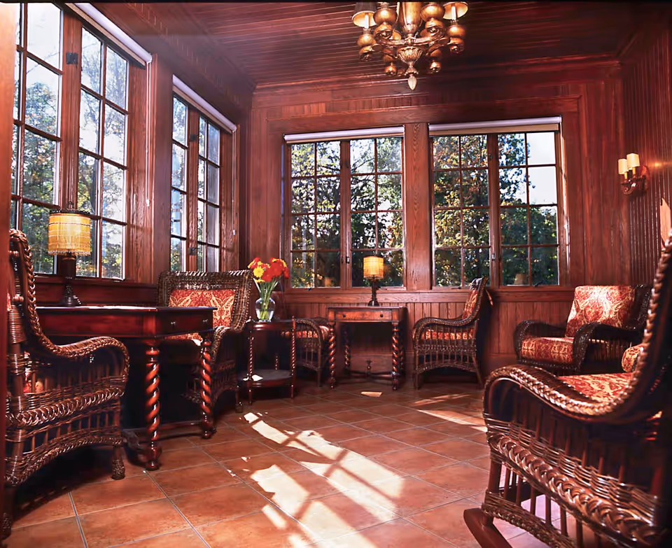A cozy sunroom with large windows letting in natural light, featuring wicker chairs with patterned cushions, wooden side tables with lamps, a small table with a vase of flowers, and wood-paneled walls and ceiling.