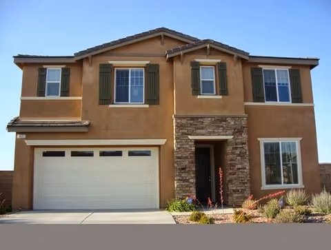 Two-story house with a beige stucco exterior, green window shutters, a stone accent around the front door, and a two-car garage. The front yard has desert landscaping with small plants and gravel.