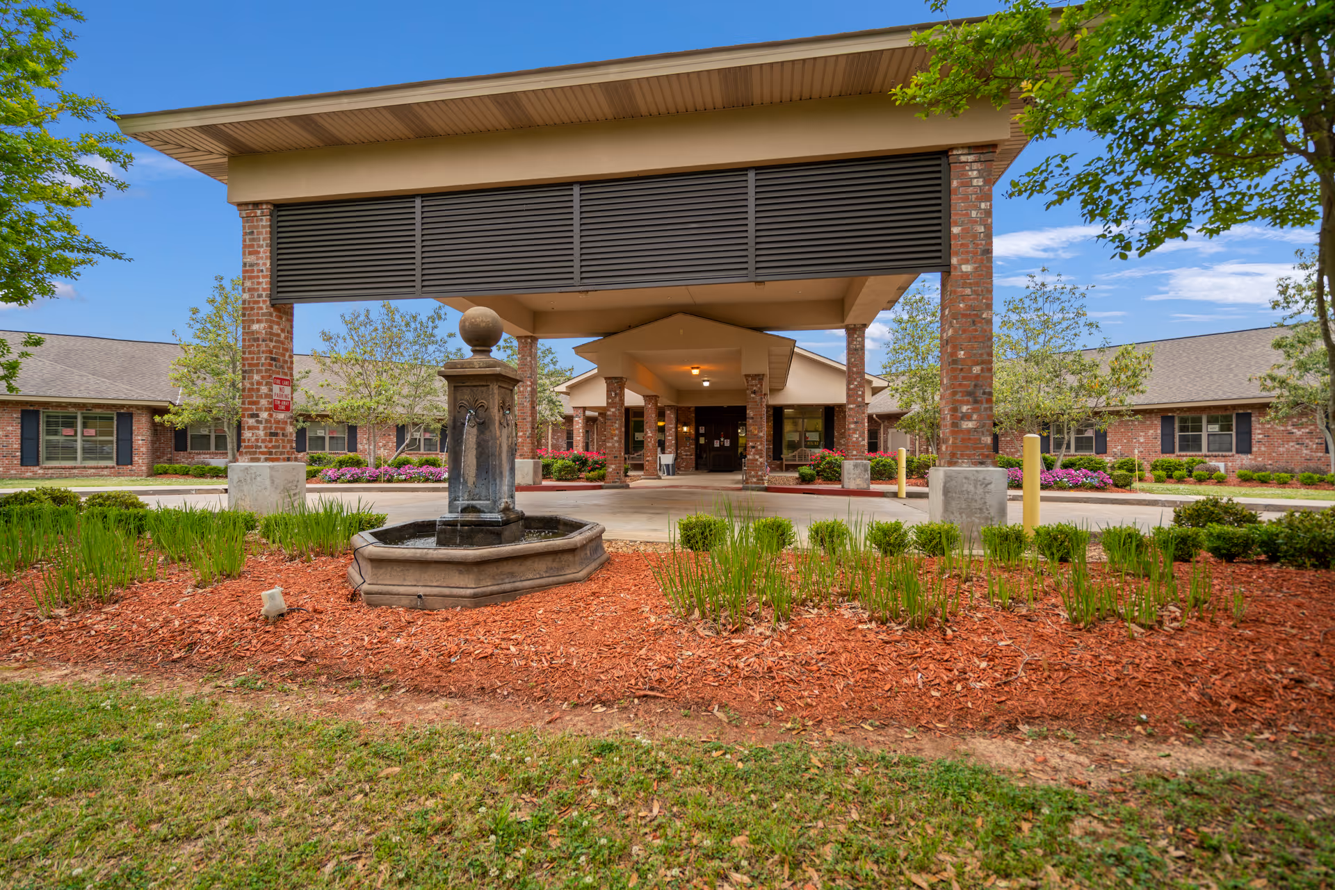 Front entrance of Heritage Manor West featuring brick columns, a covered drop-off area, and a central fountain with landscaped beds.
