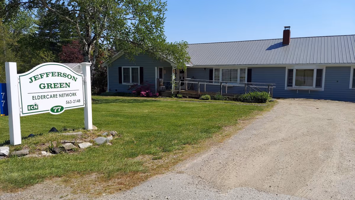 Exterior view of Jefferson Green Eldercare Network building with a green lawn, a gravel driveway, and a white sign displaying the facility name and contact information. The building is a single-story structure with blue siding, white trim, and a metal roof.